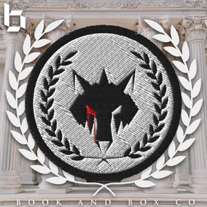 以下が含まれることがあります： 黒と白の刺繍パッチ。狼の頭のシルエットと赤い血のしずくが描かれている。パッチは月桂樹の輪と黒い円に囲まれている。