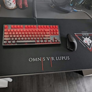 Könnte beinhalten: Eine schwarze Schreibtischunterlage mit einer rot beleuchteten mechanischen Tastatur, einer schwarzen Maus und dem Text "OMNIS VR LUPUS". Die Unterlage zeigt auch ein Wolfslogo und den Text "HOWLER EST. 7:30 PM".