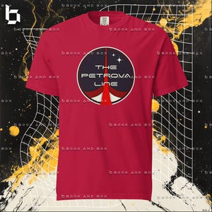 May include: A red t-shirt featuring a circular graphic with the text "THE PETROVA LINE". The graphic is a dark blue circle with a red and white rocket design. The shirt is a solid red color.