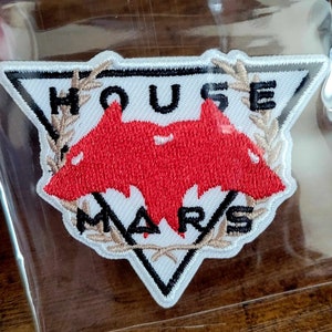 May include: White triangular patch with black stitching and a red bat-like figure in the centre. The text "HOUSE MARS" is embroidered in black and beige on the patch.