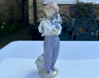 May include: A porcelain figurine of a young boy holding a small dog. The boy is wearing blue and white striped trousers and a white shirt. The dog is white with black spots. The figurine is standing on a base with a small flower pot.