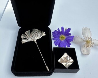 Puede incluir: Dos broches de plata en cajas de terciopelo negro. Una broche es una flor estilizada con un alfiler largo. La otra broche es una tetera, una taza y un platillo de plata en una bandeja de plata.
