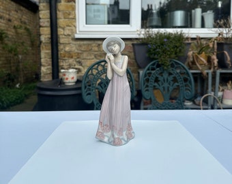 May include: A porcelain figurine of a girl wearing a white hat and a pink dress with floral details. The figurine is standing on a white surface.