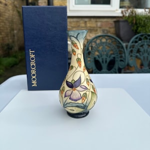 May include: A small, cream-colored ceramic vase with a blue rim and a floral design featuring purple flowers, green leaves, and orange berries. The vase is sitting on a white surface and has the word "MOORCROFT" printed on a blue box behind it.