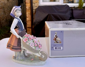 May include: A porcelain figurine of a girl in a blue and white dress pushing a wheelbarrow full of pink and white flowers. The figurine is on a white base and is next to a box with the number 01419 on it.