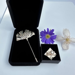 May include: Two silver brooches in black velvet boxes. One brooch is a stylized flower with a long pin. The other brooch is a silver teapot, cup, and saucer on a silver tray.