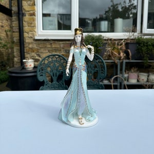 May include: A porcelain figurine of a woman wearing a blue and gold gown with a crown. She is standing on a white base.