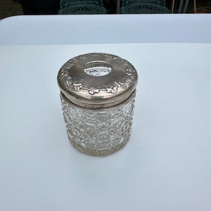 Puede incluir: Un tarro de vidrio transparente con una tapa de plata. La tapa tiene un diseño decorativo de hojas y flores. El tarro tiene un patrón texturizado.