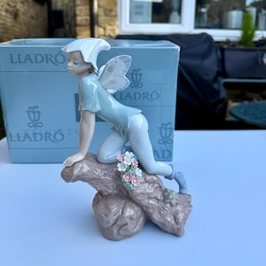 May include: A porcelain figurine of a fairy with blue wings and a white hat, perched on a tree branch with pink and white flowers. The figurine is made by Lladró.