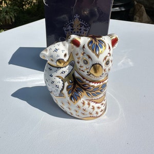 May include: A white porcelain figurine of a mother koala and her joey. The mother koala is sitting upright with her joey clinging to her back. Both koalas are decorated with intricate blue, gold, and red patterns.