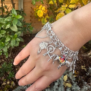 May include: A silver charm bracelet with a variety of charms, including a small house, a wheel, a horse and rider, and a bird. The bracelet is on a person's wrist.