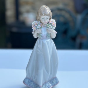 May include: A porcelain figurine of a girl in a white dress holding two bouquets of pink and white flowers.