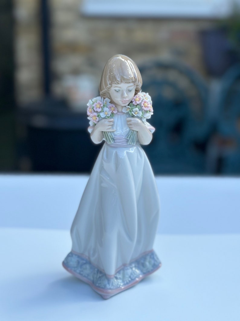 May include: A porcelain figurine of a girl in a white dress holding two bouquets of pink and white flowers.