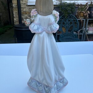 May include: A porcelain figurine of a girl wearing a white dress with a blue and pink floral pattern. She has long brown hair and is holding a bouquet of pink flowers.