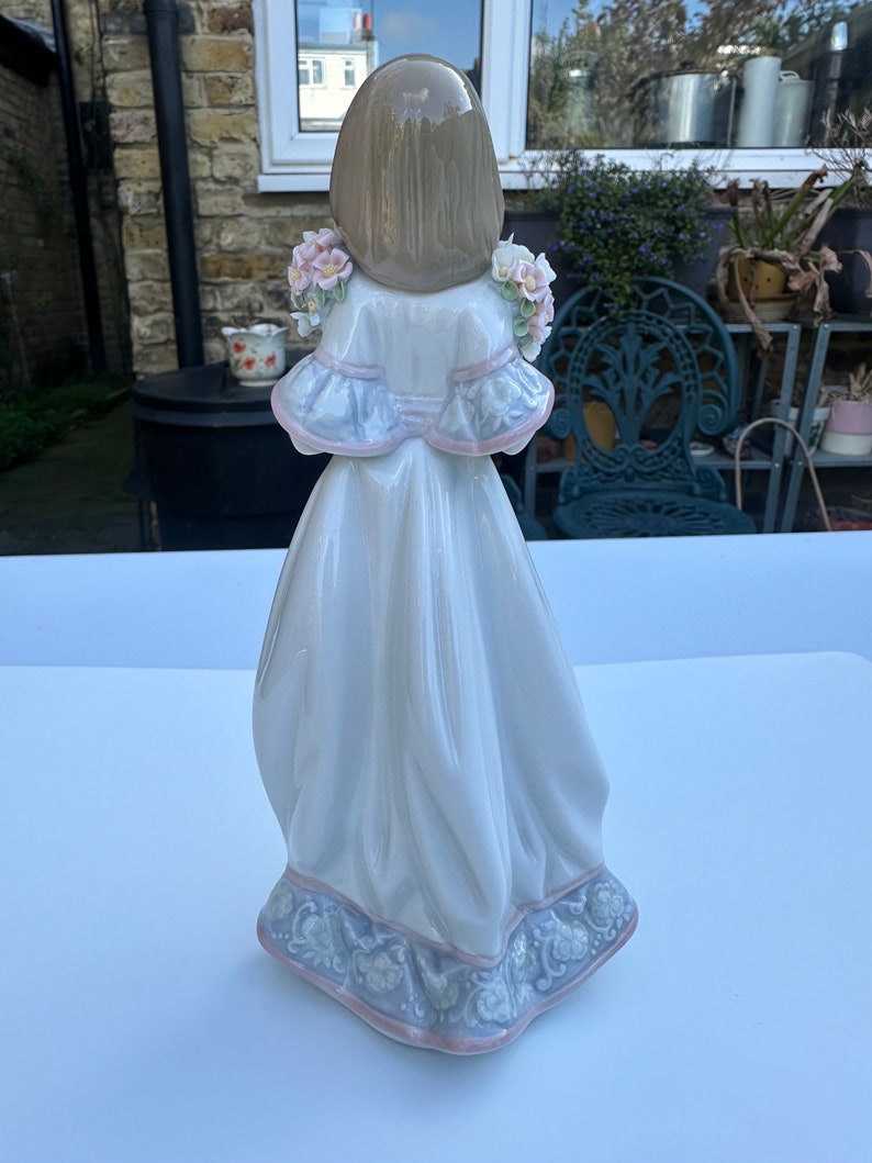 May include: A porcelain figurine of a girl wearing a white dress with a blue and pink floral pattern. She has long brown hair and is holding a bouquet of pink flowers.