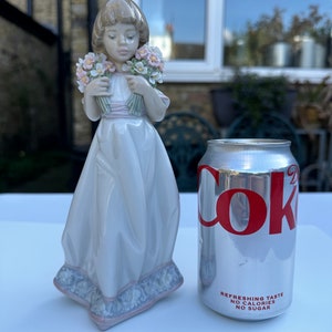 May include: A porcelain figurine of a girl holding two bouquets of pink and white flowers. She is wearing a white dress with a pink sash and a blue and white patterned base. The figurine is next to a can of Coca-Cola Zero Sugar.