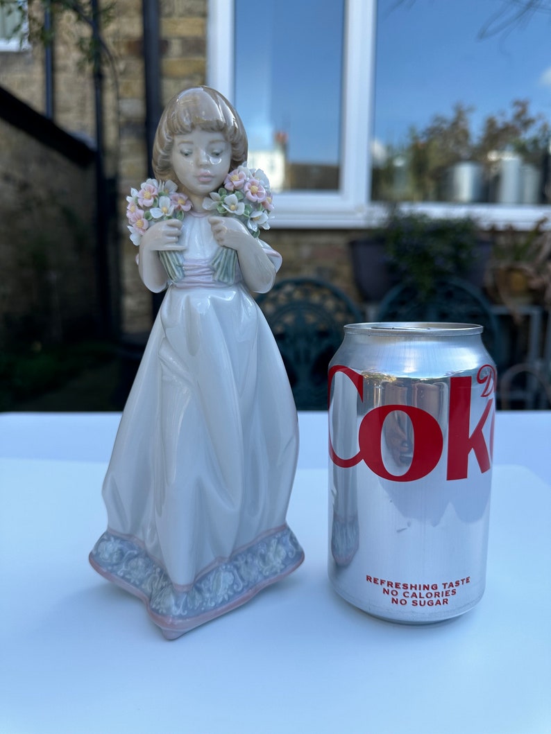 May include: A porcelain figurine of a girl holding two bouquets of pink and white flowers. She is wearing a white dress with a pink sash and a blue and white patterned base. The figurine is next to a can of Coca-Cola Zero Sugar.