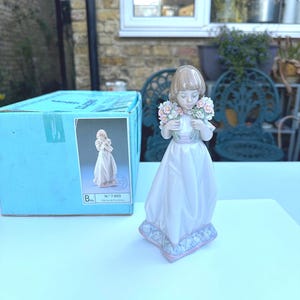 May include: A porcelain figurine of a girl holding a bouquet of flowers. She is wearing a white dress with a pink sash and has brown hair. The figurine is on a blue and white base. The box reads "N". 7,603.