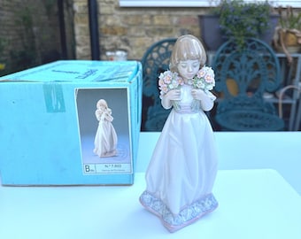 May include: A porcelain figurine of a girl holding a bouquet of flowers. She is wearing a white dress with a pink sash and has brown hair. The figurine is on a blue and white base. The box reads "N". 7,603.