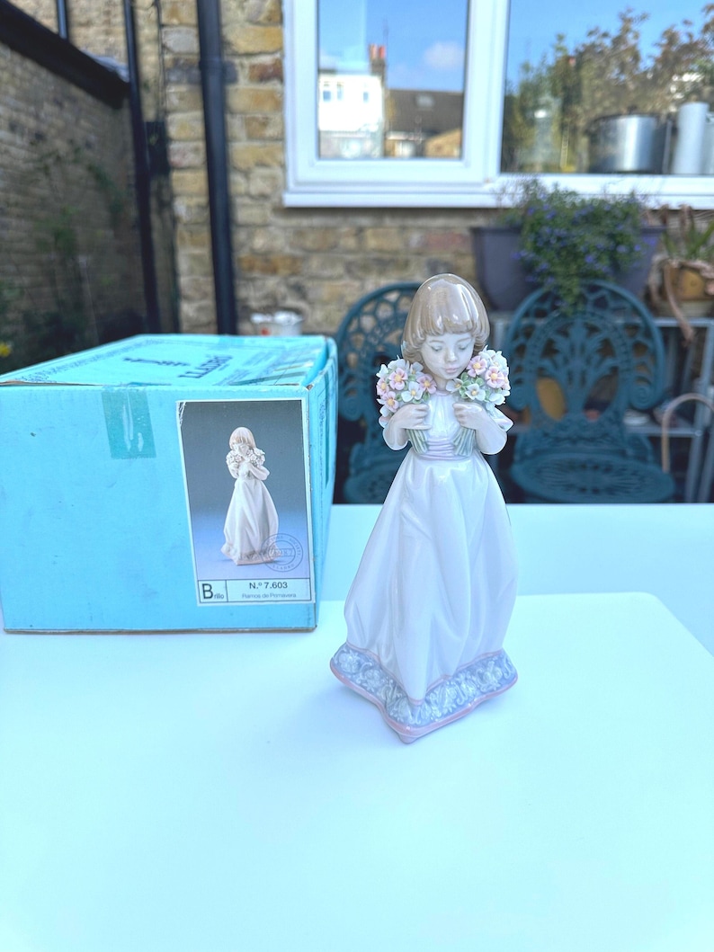 May include: A porcelain figurine of a girl holding a bouquet of flowers. She is wearing a white dress with a pink sash and has brown hair. The figurine is on a blue and white base. The box reads "N". 7,603.