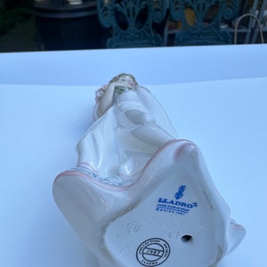 May include: A porcelain figurine of a girl holding a bouquet of flowers. The figurine is wearing a white dress and has a pink sash. The base of the figurine is marked with the Lladro logo and the year 1987.