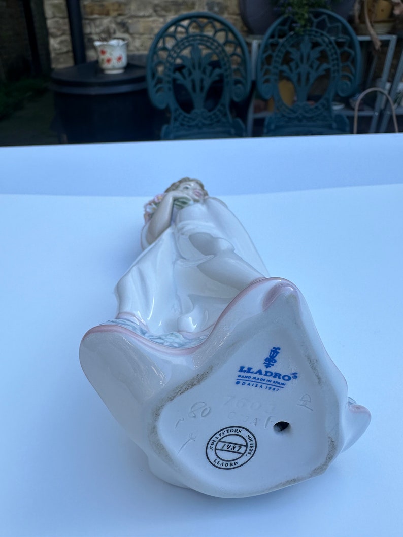 May include: A porcelain figurine of a girl holding a bouquet of flowers. The figurine is wearing a white dress and has a pink sash. The base of the figurine is marked with the Lladro logo and the year 1987.