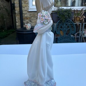 May include: A porcelain figurine of a girl in a white dress holding a bouquet of pink and white flowers. The figurine is standing on a white surface.