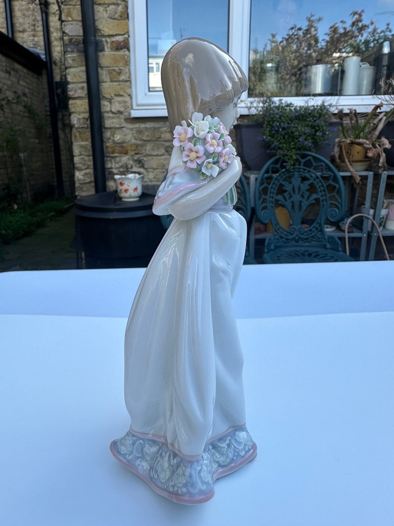 May include: A porcelain figurine of a girl in a white dress holding a bouquet of pink and white flowers. The figurine is standing on a white surface.
