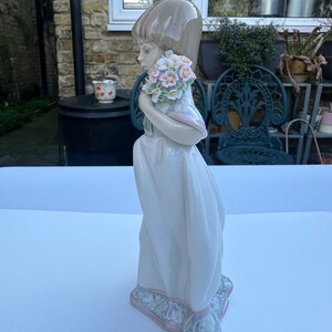 May include: A porcelain figurine of a girl in a white dress holding a bouquet of pink and white flowers. The figurine is standing on a white surface.
