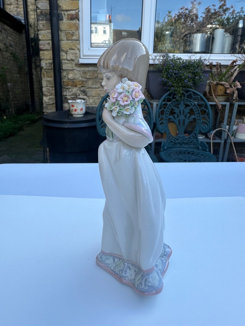 May include: A porcelain figurine of a girl in a white dress holding a bouquet of pink and white flowers. The figurine is standing on a white surface.