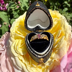 May include: A gold ring with a central ruby and diamond accents, displayed in a heart-shaped black jewelry box. The box is open, revealing the ring against a black velvet interior. The background features a yellow and pink rose.