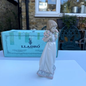 May include: A porcelain figurine of a girl in a white dress holding a bouquet of pink and white flowers. The figurine is standing on a blue and white base. The figurine is made by Lladr&oacute;.