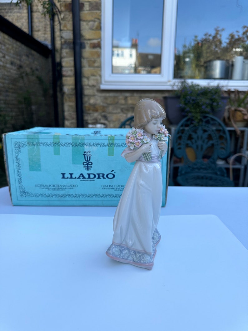 May include: A porcelain figurine of a girl in a white dress holding a bouquet of pink and white flowers. The figurine is standing on a blue and white base. The figurine is made by Lladr&oacute;.