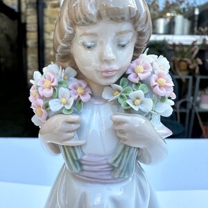 May include: A porcelain figurine of a girl holding two bouquets of pastel pink, white, and green flowers. The girl is wearing a white dress with a pink sash.