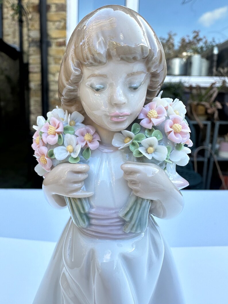 May include: A porcelain figurine of a girl holding two bouquets of pastel pink, white, and green flowers. The girl is wearing a white dress with a pink sash.