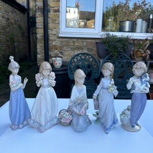 May include: Set of five porcelain figurines of girls in various poses, each holding flowers or a small animal. The figurines are painted in pastel colors and have delicate details.