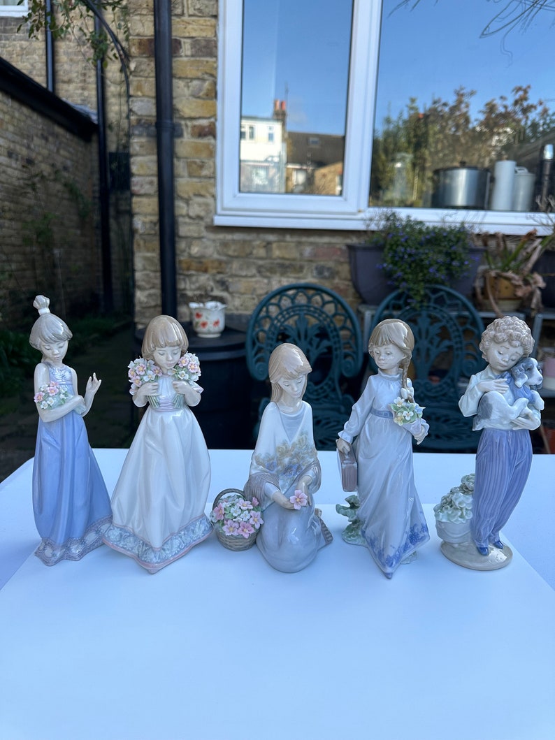 May include: Set of five porcelain figurines of girls in various poses, each holding flowers or a small animal. The figurines are painted in pastel colors and have delicate details.