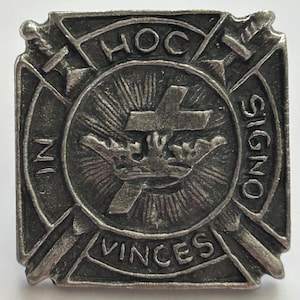 Könnte beinhalten: Quadratische, silberfarbene Metallbrosche mit zentralem kreisförmigen Design. Sie zeigt ein Kreuz, eine Krone und die Worte "HOC SIGNO VINCES." Vier Schwerter umgeben den Kreis, mit den Worten "IN" und "HOC" auf beiden Seiten.