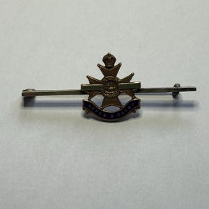 Könnte beinhalten: Eine goldfarbene Anstecknadel mit einem blauen und grünen Emaille-Design. Die Nadel zeigt ein Kreuz mit einer Krone oben und eine Schriftrolle mit dem Text "NOTTO & DERBY".