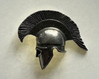 May include: A silver-colored metal pin shaped like a Greek warrior helmet with a plume on top.