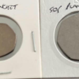 May include: Two heptagonal metal coin blanks in white cardboard holders. The left blank is copper-coloured with the handwritten text 20p Planchot above it. The right blank is silver-coloured with the handwritten text 50p Planchot above it.