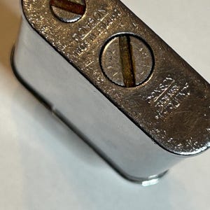 May include: A chrome-plated Ronson lighter with visible screws and the text "RONSON MADE IN ENGLAND". The lighter has a rectangular shape with rounded edges and a shiny, reflective surface. The lighter is a vintage collectible.