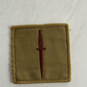 以下が含まれることがあります： 中央にダークブラウンの短剣が刺繍された、ベージュ色の正方形の布製ワッペン。ワッペンには同系色のベージュの糸で縁取りが施されており、正方形のテキスタイルデザインの周囲をすっきりと仕上げています。