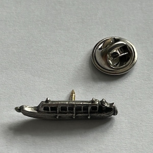 May include: A silver-toned metal pin of a narrow boat with a pointed bow and stern. The pin has a gold-coloured pin back.