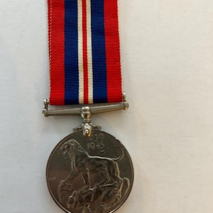 Puede incluir: Una medalla de plata con un león de pie sobre una montaña y el texto "1939 1945" en el anverso. La medalla está unida a una cinta roja, blanca y azul.