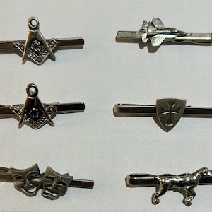 May include: Six silver tie clips with different designs. The designs include a compass and square, a jet fighter, a cross in a shield, two theater masks, and a dog.