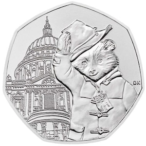 Könnte beinhalten: Eine silberne Gedenkmünze mit Paddington Bear, der einen blauen Mantel und einen Hut trägt und vor der St. Paul's Cathedral in London steht. Die Münze trägt den Text "Paddington Bear" und "The London 2022 50p Coin".