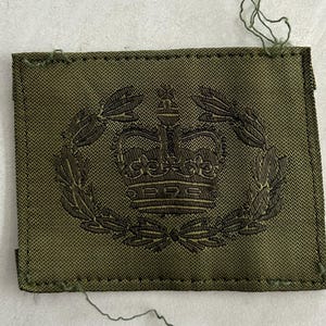 May include: A square olive green fabric patch featuring a dark green embroidered design of a crown centered within a circular laurel wreath. The patch has visible stitching along the edges and loose threads at the top and bottom corners.