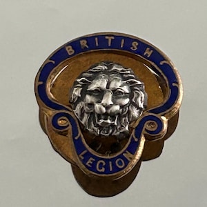 May include: A circular metal pin featuring a silver-toned lion head at the centre. The lion is surrounded by a blue enamel border with the gold-coloured text BRITISH LEGION inscribed along the top and bottom of the circular frame.