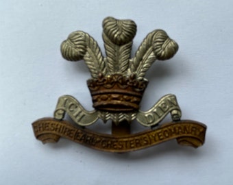 Puede incluir: Un distintivo de gorra militar dorado y plateado con una corona y tres plumas. El texto en el rollo dice "Cheshire Earl Chester's Yeomanry ICH DIEN".
